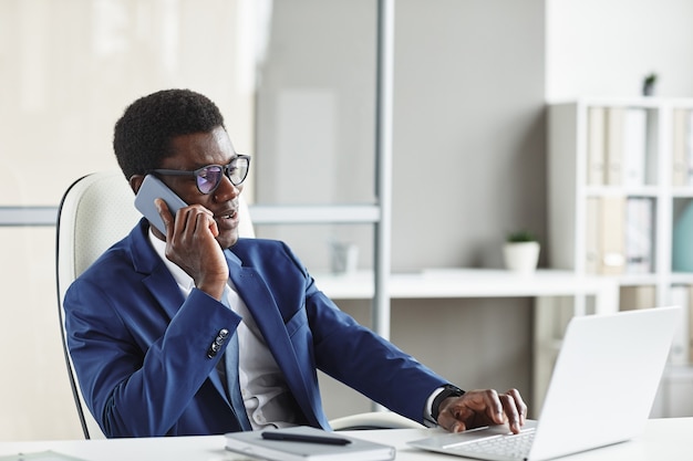 Empresário africano usando óculos fazendo seu trabalho no laptop enquanto fala ao telefone em seu local de trabalho no escritório