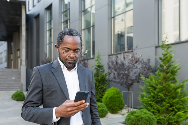 Empresário afro-americano sério e bem-sucedido fora do prédio de escritórios usando smartphone