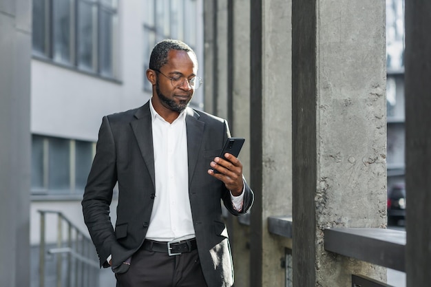 Empresário afro-americano sério e bem-sucedido fora do prédio de escritórios usando smartphone