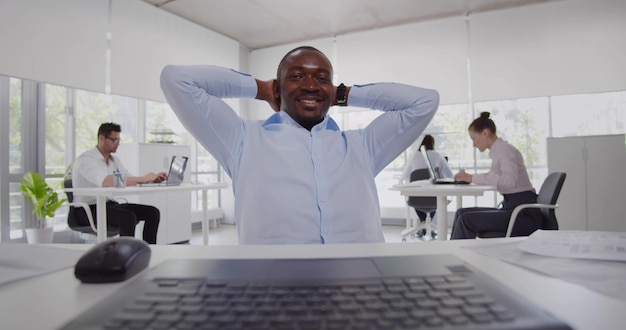 Empresário afro-americano sorridente segurando as mãos atrás da cabeça sentado na mesa do escritório