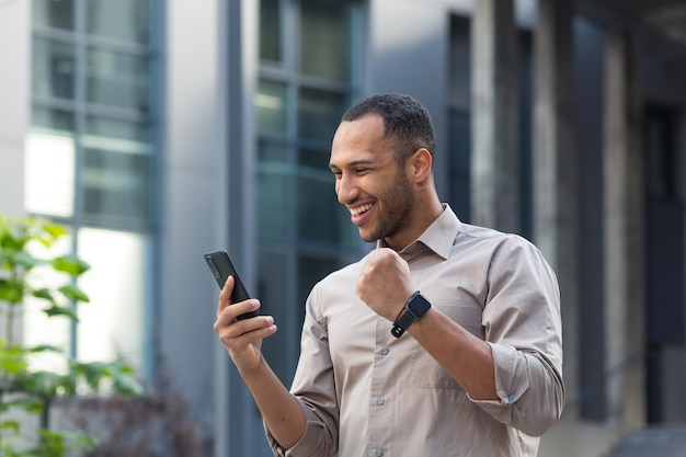 Empresario afroamericano fuera del edificio de oficinas usando el teléfono sonriendo y feliz levantando la mano