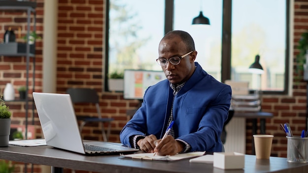 Empresario afroamericano tomando notas en una laptop, usando estadísticas de Internet para crear una presentación de inicio. Trabajador de la empresa escribiendo correo electrónico, navegando por el sitio web para planificar el proyecto. Disparo de trípode.