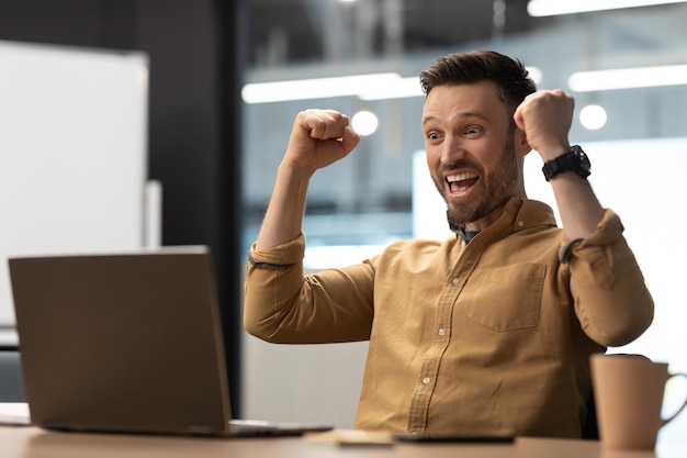 Empresário alegre usando laptop e agitando os punhos sentado no escritório