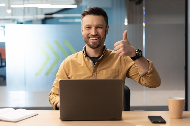 Empresário alegre usando laptop e gesticulando polegares para cima no escritório