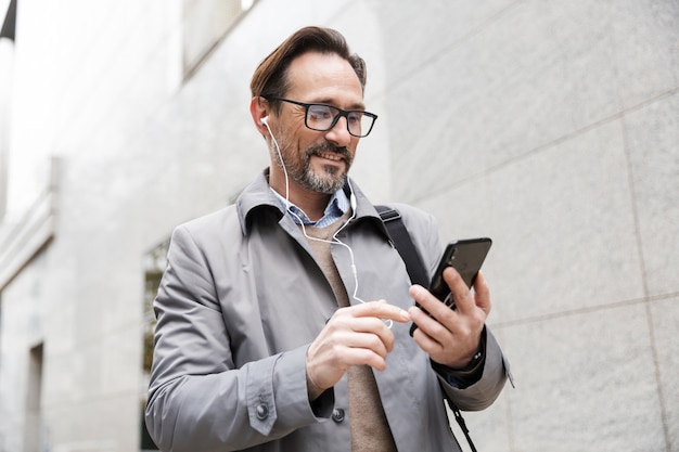 empresário alegre usando óculos, usando celular e fones de ouvido, perto de um prédio comercial