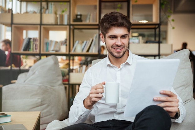 Empresário alegre vestido de camisa branca, sentado no café e lendo documentos enquanto bebia chá. Olhando documentos.