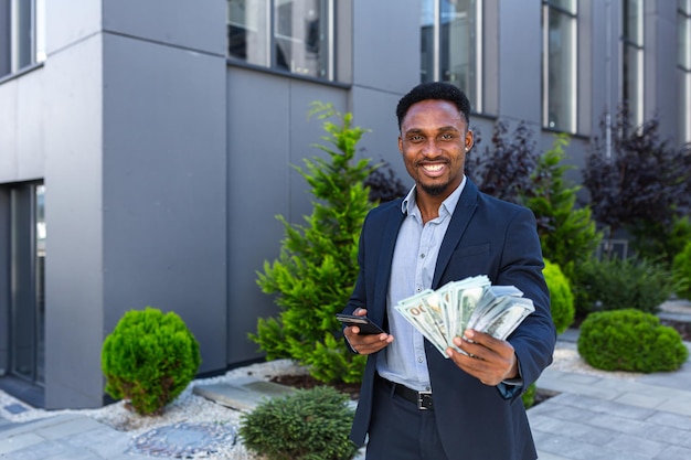 Empresário americano africano em pé na rua edifício moderno mostra ganhos ganhando em apostar dinheiro em espécie com o telefone móvel nas mãos. homem negro de terno ao ar livre com smartphone e pacote de dólares