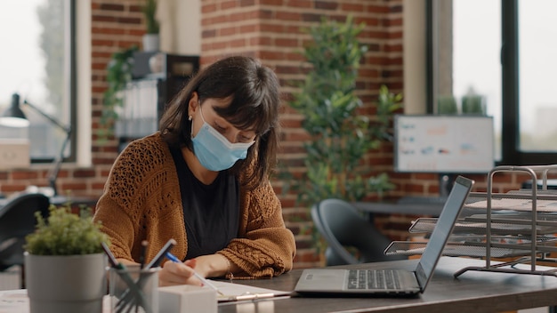 Empresario analizando archivos de gráficos y pantalla de computadora portátil en el escritorio para planificar la estrategia comercial y el proyecto. Mujer que trabaja con análisis e información, usando mascarilla durante la pandemia