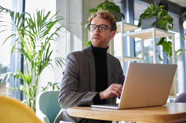 Empresario con anteojos elegantes y traje usando una computadora portátil que trabaja en línea en el lugar de trabajo