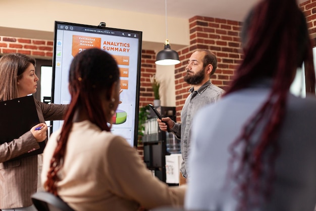 Empresario apuntando al monitor explicando las estadísticas de marketing al equipo de negocios que trabaja en la presentación de la empresa en la oficina de inicio. Diversos empresarios planificando presentaciones comerciales.