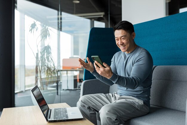 Empresário asiático bem-sucedido comemora vitória olha para o telefone feliz e se alegra sentado no escritório moderno