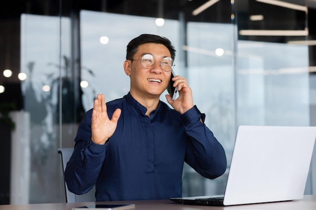 Empresário asiático bem-sucedido e alegre falando ao telefone com clientes dentro do escritório maduro