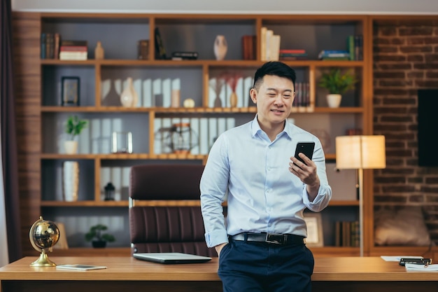 Empresário asiático bem-sucedido no escritório doméstico moderno olha para os sorrisos da tela do telefone e se alegra com o retrato do freelancer de sucesso na camisa usa o aplicativo de telefone