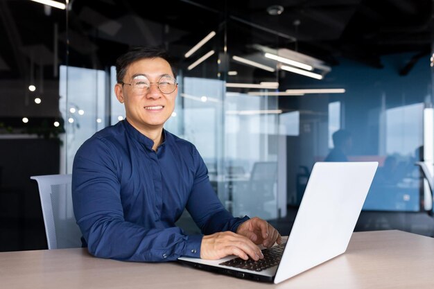 Empresário asiático bem-sucedido sorrindo e olhando para o programador de câmera dentro do escritório usando