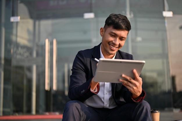 Empresário asiático bonito usando seu tablet enquanto está sentado na escada do lado de fora do prédio da empresa