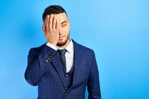 Empresário asiático cazaque de terno azul com gravata faz gesto de palma do rosto isolado no fundo do estúdio. Fracasso, perdedor, dói assistir o conceito