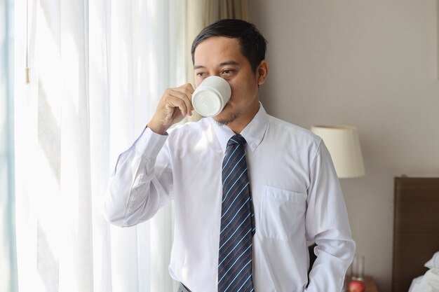 Empresário asiático de camisa branca e gravata em pé enquanto bebe uma xícara de café em casa