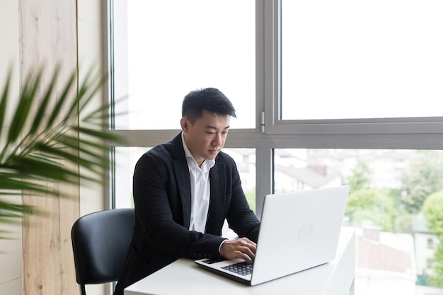 Empresário asiático de sucesso em um terno preto trabalha em um laptop em um escritório elegante