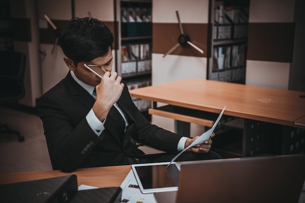 Empresário asiático de terno preto pensa em ideia para trabalhar no escritório ele trabalha horas extras sozinho em uma empresa de tons escuros