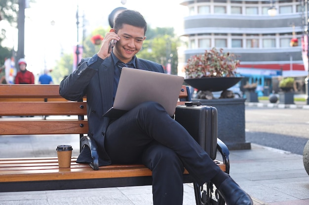 Empresário asiático de terno sentado no banco enquanto abre o laptop e faz uma ligação