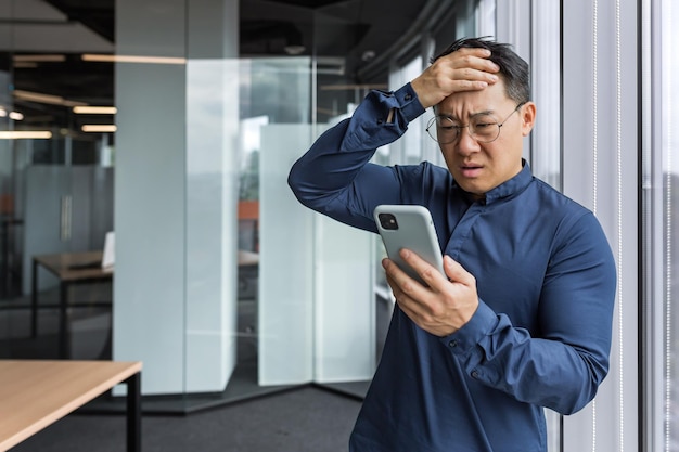 Empresário asiático dentro do prédio de escritórios chateado e triste lendo más notícias no negócio de telefonia móvel