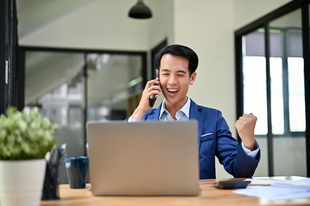 Empresário asiático entusiasmado mostrando um punho cerrado recebendo boas notícias por telefone