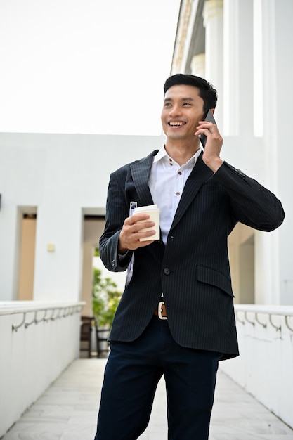 Empresário asiático inteligente em traje formal do lado de fora do prédio e falando ao telefone