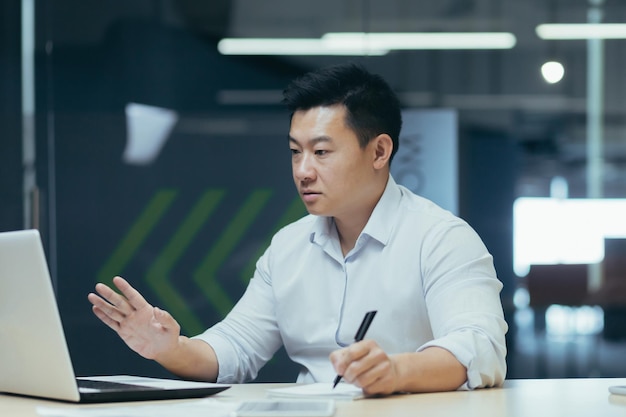 Empresario asiático preocupado sentado en la mesa de la oficina con una laptop hablando en una videollamada
