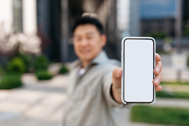 Empresario asiático que muestra un teléfono inteligente con pantalla en blanco a la cámara posando al aire libre cerca de la maqueta del edificio de oficinas