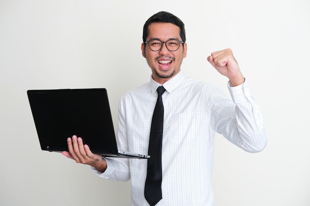 Foto empresário asiático segurando um laptop enquanto punho cerrado e mostrando expressão confiante