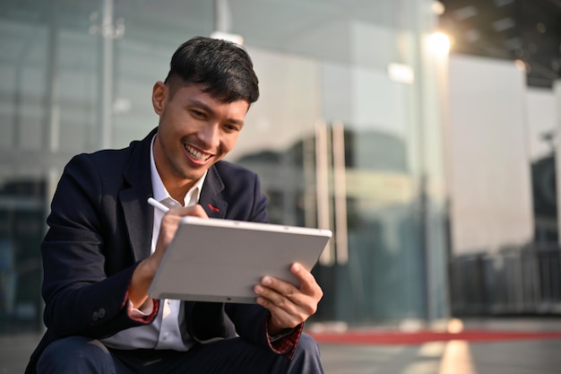 Empresario asiático sonriente usando su tableta mientras se sienta en las escaleras fuera del edificio de la empresa