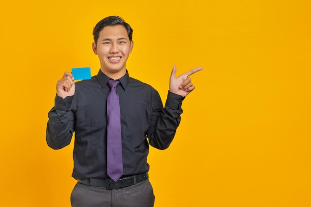 Empresário asiático sorridente segurando um cartão de crédito enquanto aponta o dedo para o espaço da cópia em amarelo