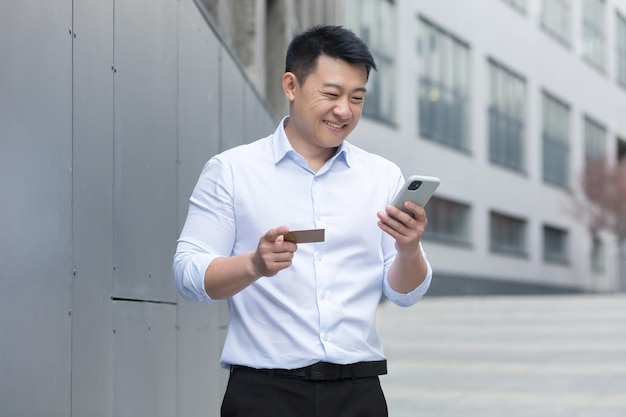 Empresário asiático sorrindo e feliz fazendo compras online fora do prédio de escritórios usando celular