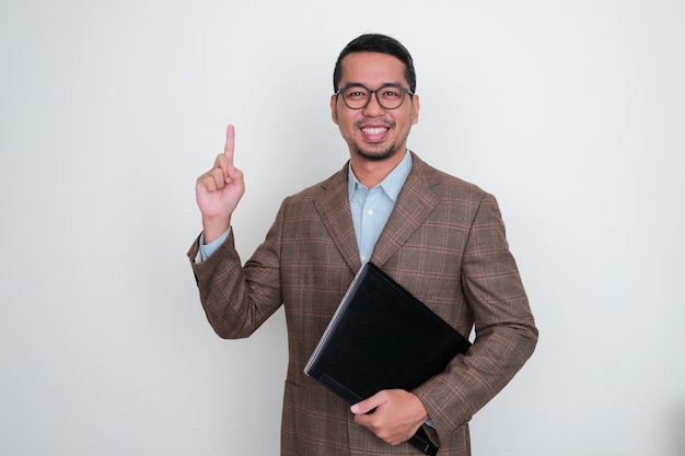 Empresario asiático con traje marrón sonriendo y sosteniendo una laptop apuntando con el dedo hacia arriba