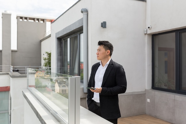 Empresario asiático en traje de negocios disfruta de teléfono de pie en el balcón terraza del moderno centro de oficinas