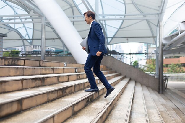 Empresário asiático vestindo paletó correndo rápido lá em cima vai trabalhar de manhã ao ar livre.