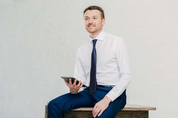 Empresário atraente pensativo pensa em uma nova ideia criativa usa touch pad moderno vestido elegantemente senta-se na mesa de madeira sozinho posa contra a parede branca Prospero financista masculino interior