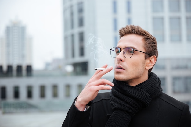 Empresário bacana de óculos e roupas quentes fumando cigarro na rua