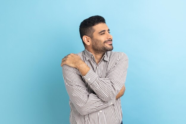 Empresario con barba de pie y abrazándose a sí mismo con una sonrisa dentuda disfrutando de una autoestima positiva