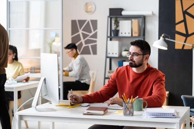 Foto empresario barbudo con anteojos sentado en la mesa y mirando el monitor de la computadora que trabaja en la oficina con sus colegas en el fondo