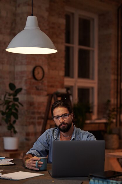 Foto empresário barbudo bebendo café e olhando para o monitor do laptop enquanto trabalhava na mesa do escritório