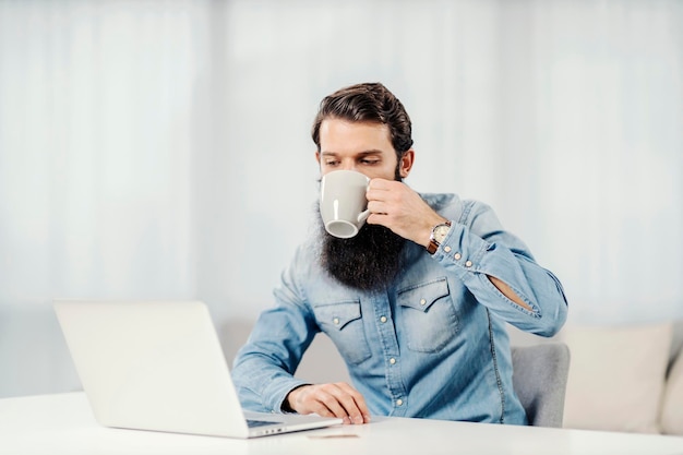 Un empresario barbudo bebiendo su café y mirando una laptop en casa