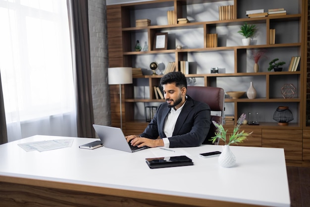 Empresário barbudo bem-sucedido em traje elegante trabalhando em laptop em escritório moderno