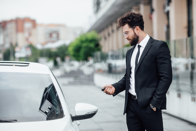 Empresário barbudo bonito em terno preto entrando em seu carro enquanto fica ao ar livre nas ruas da cidade perto do moderno centro de escritórios