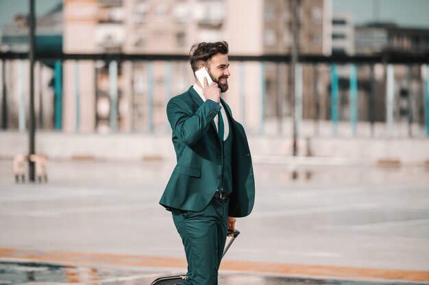 Empresário barbudo caucasiano ambicioso no terno usando telefone inteligente e carregando bagagem enquanto acorda no estacionamento. Conceito de viagem de negócios. Não olhe para trás, você não vai por esse caminho.