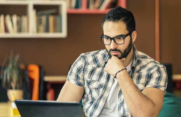 Empresário barbudo com óculos trabalhando com laptop na moderna sala de coworking elegante com pensamento inteligente de estantes