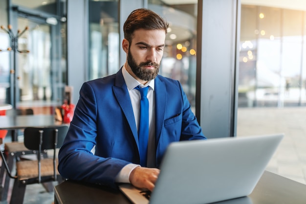 Empresário barbudo com roupa formal com expressão facial séria usando laptop enquanto está sentado na lanchonete ao lado da janela.