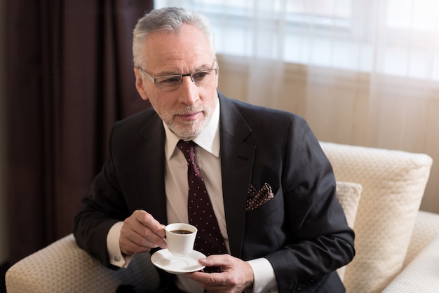 Empresário barbudo envelhecido e encantado segurando uma xícara e bebendo café enquanto está sentado no hotel