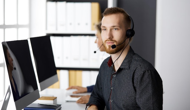 Empresário barbudo falando pelo fone de ouvido perto de sua colega enquanto está sentado no escritório moderno. Grupo de pessoas diversas no call center. Telemarketing e atendimento ao cliente.