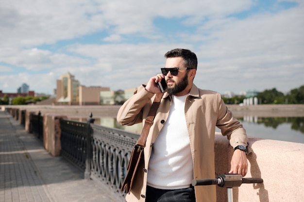 Empresario barbudo en gabardina beige, jersey de punto blanco y gafas de sol hablando por teléfono inteligente mientras está de pie junto al río